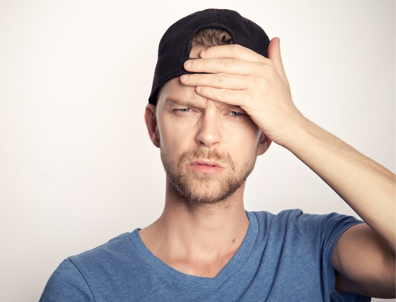 A frustrated man in a cap and blue shirt holding his forehead.