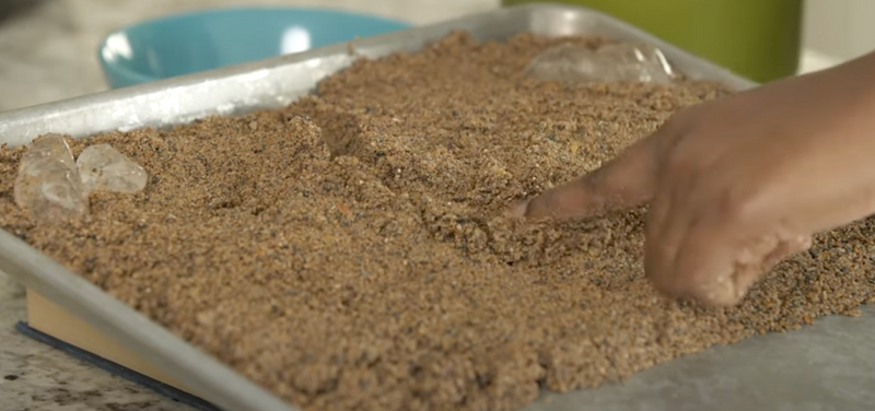 Tray with sand and ice cubes on both upper corners. A woman traces her two fingers down the sand to create a riverbed.