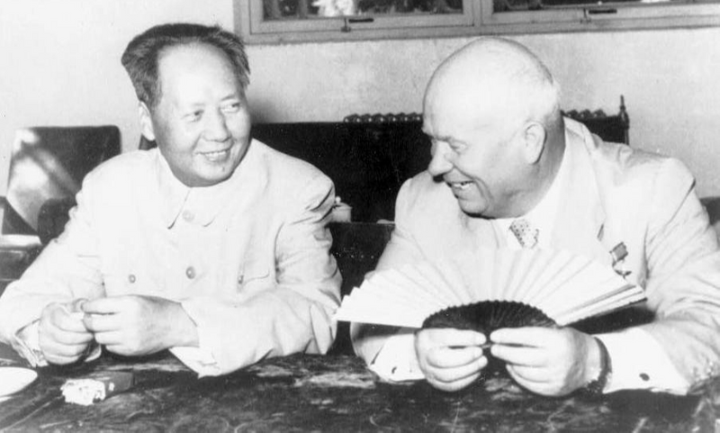 Mao Tsetung and Nikolai Kruschev at a meeting. They hold Chinese paper fans in their hands and smile at each other.