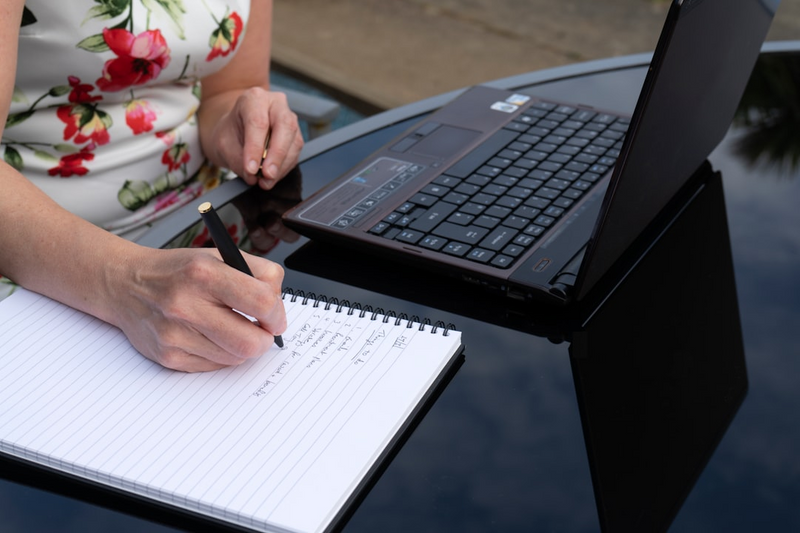 A woman seated at a table with her laptop, writing a list in a notebook.