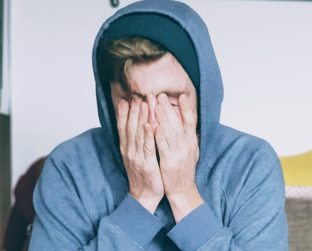 A young man wearing a hoodie, sitting on a couch covering his face with his hands.