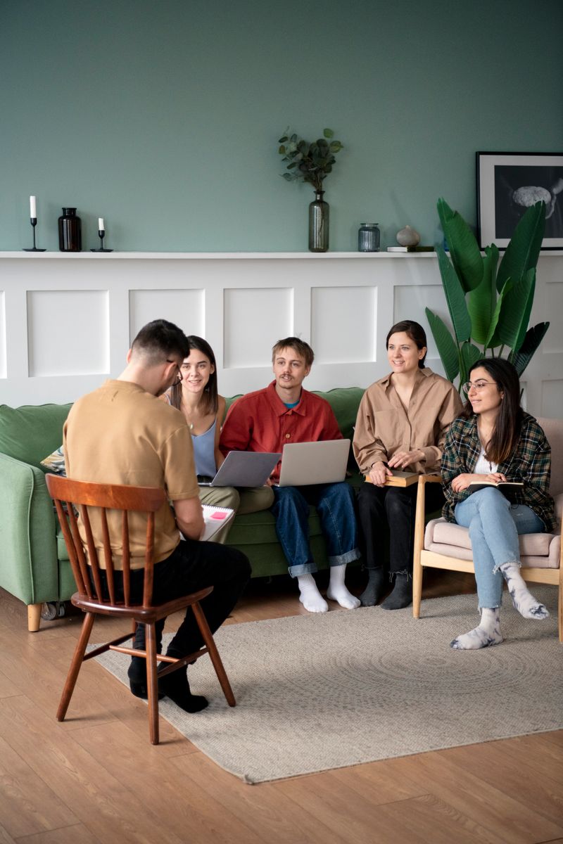 Five adults sitting together in a living room, facing one person who is seated on a chair and holding a notebook.