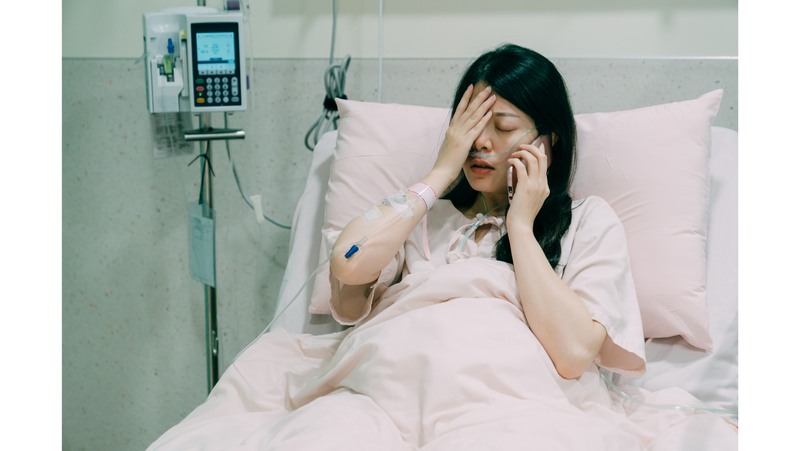 A woman reclines in a hospital bed. One hand is on her forehead, the other holding a phone to her ear. She looks strained.