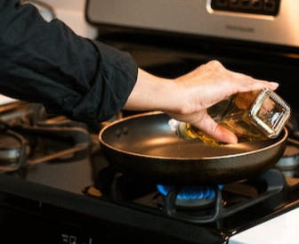 Woman's black-sleeved hand pouring cooking oil into a frying pan on a gas heated stovetop.