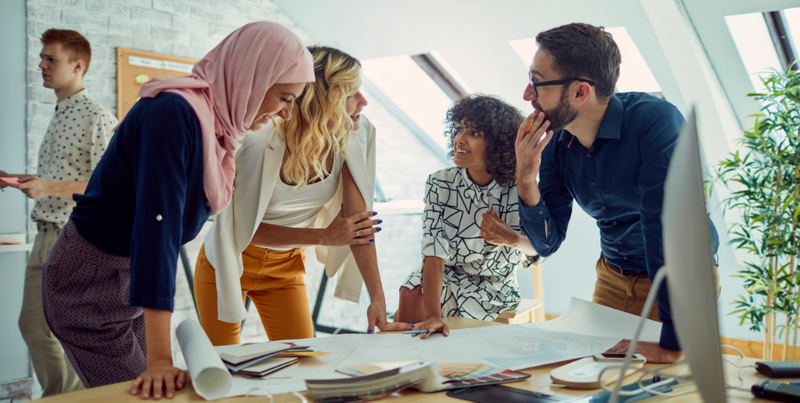 A group of people at work from diverse backgrounds standing and discussing a project.  