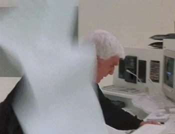 GIF: Man with short white hair frantically watches paper fly out of the printer from an old computer.