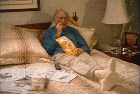 An old man snacks on chips while resting on the bed.