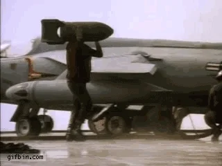 Two Air Force technicians carry bombs on a flight deck. They bump into each other.