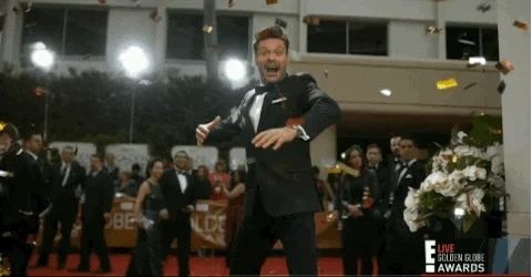 Broadcaster Ryan Seacrest on red carpet at awards show with excited face, making wide arm gesture while gold confetti flies