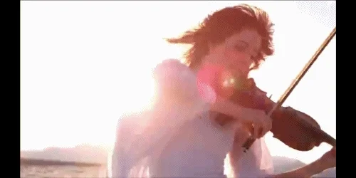 Violinist Lindsey Stirling wearing a white flowing dress and playing the violin outside with sun in the background.