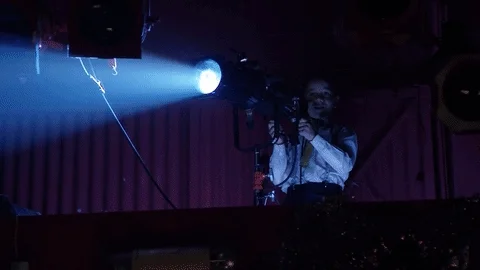 A bright stage light shines on a scared looking man on stage.