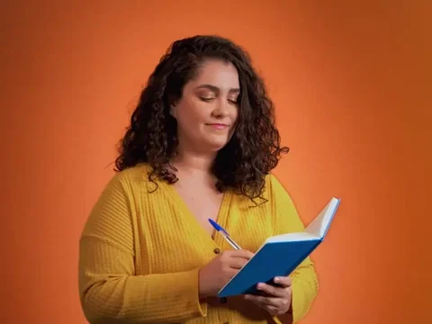 A woman with long, curly hair and a yellow top nodding in agreement as she prepares to write in a notebook.