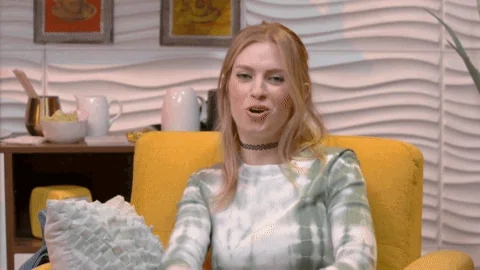 A blonde woman sitting in a mustard yellow chair, gesturing to the right with a finger gun. She says, 