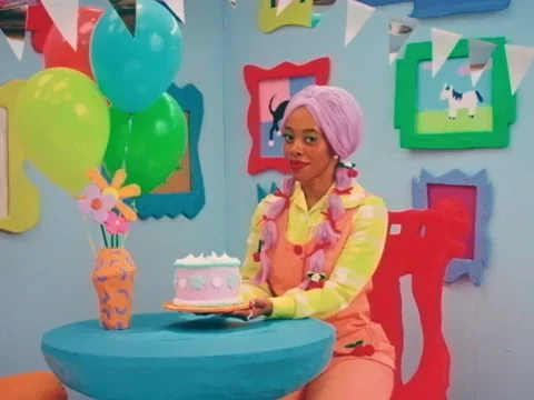 Person in colorful room wearing a lavender wig with braids. They're holding a cake and the text says 