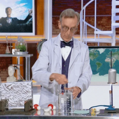 Bill Nye in a lab. He stirs a mixture in a beaker.