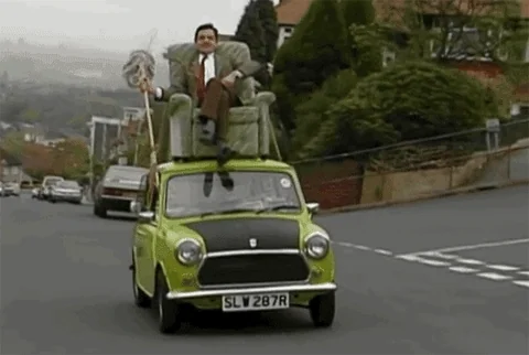 Mr. Bean sitting on the roof of a moving car. He uses a mop contraption to steer.