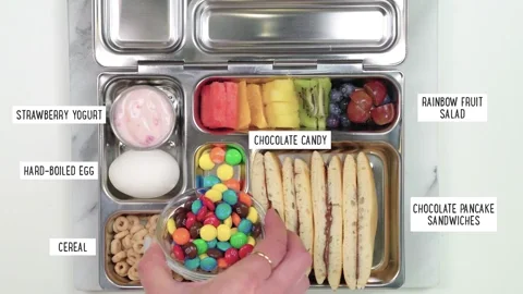 A metal lunch box. It contains yogurt, an egg, cereal, chocolate pancake sandwiches, rainbow fruit salad, and candy.