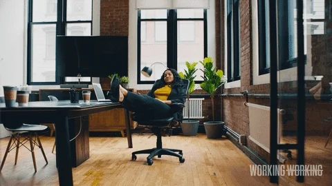 A woman grins as she leans back in her chair with her feet up on the desk as she folds her arms behind her head. 