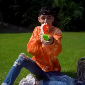 a boy kneeling on a grassy lawn uses a large water gun and sprays the camera