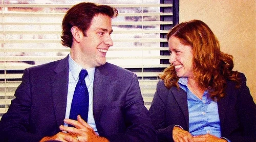 Two people sitting next to each other. They look at each other and turn away laughing.
