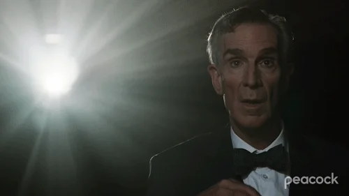 Bill Nye eating popcorn in a cinema. A project light shines behind him.