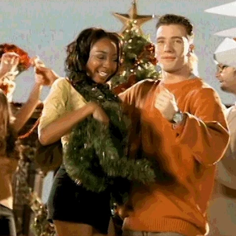 People decorated with tinsel and Santa hats signing and dancing between a Christmas tree and sleigh.