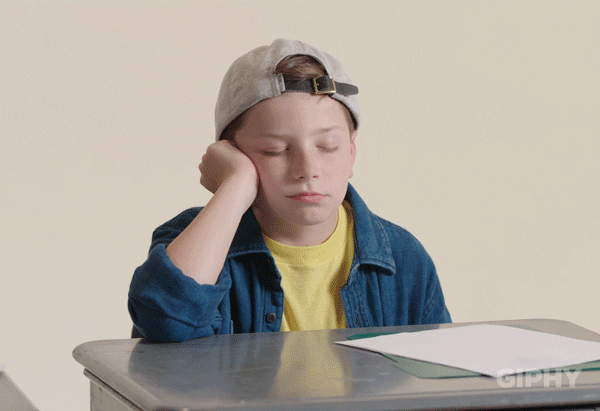 Child sitting at a desk with papers on it looking bored. Starts to fall asleep and jolts himself awake again.