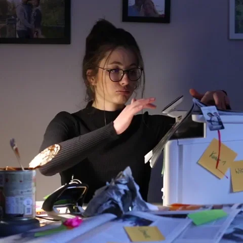 A  woman with brown hair and glasses sitting at an office desk surrounded by piles of paperwork.