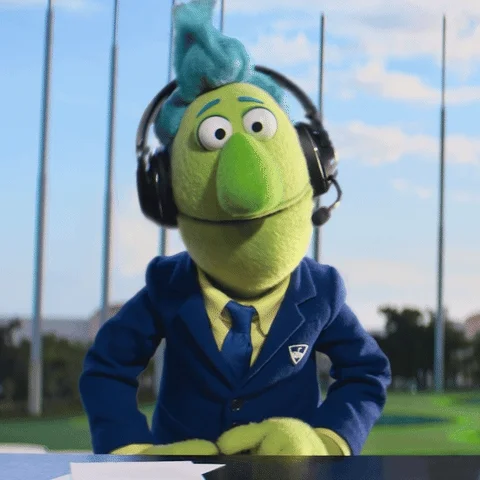 A puppet dressed up as a TV commentator, animatedly speaking and pointing at the camera. 