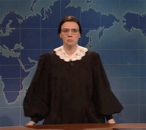 Kate McKinnon has on a brown wig and glass. She is shimmying while wearing a black judge's robe and a lace collar.