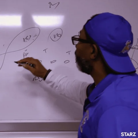 Man wearing blue shirt and cap, writing a game plan with a marker on a white board.
