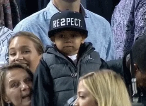 A small boy in a crowd. He wears a hat that with a logo that reads 
