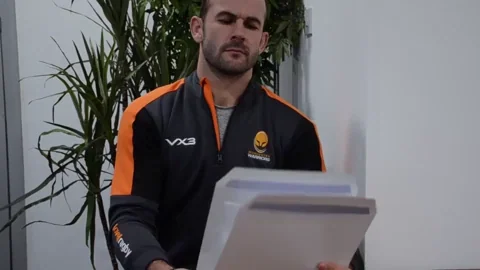 A man , slightly bald, wearing a black and orange sweatshirt, is stifling a bunch of papers and giving a disgusted look.