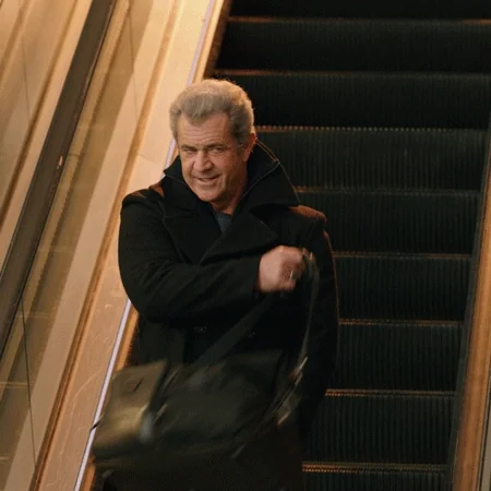 Mel Gibson on an escalator swinging his bag over his shoulder, looking confident and ready.