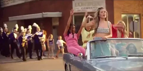 A group of young women happily drive down a street in a convertible. A marching band follows them.