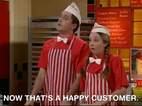 Two people dressed in cafeteria server uniforms laughing. One says, 