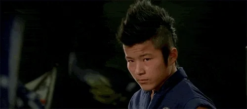 A young man with a spiky mohawk in a karate gi glares intently at the camera, nodding slightly.