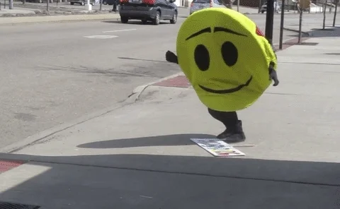 Person on siidewalk by busy stree. They're wearing a happy face costume and slowly falling over onto their back.