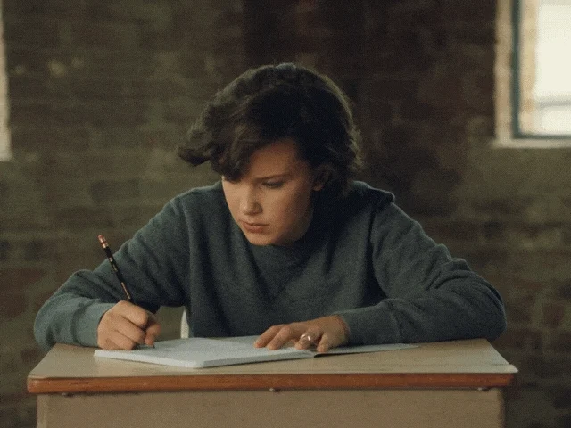 A girl is writing at a desk. She appears shocked and looks at the camera, which zooms in on her face. 