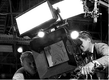 Two men with a large camera, lighting, and microphone filming a woman laying on the ground.