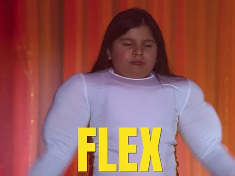 School-aged child with long, dark hair, wearing a white long-sleeved shirt, flexing their muscles, with the caption 'Flex.'