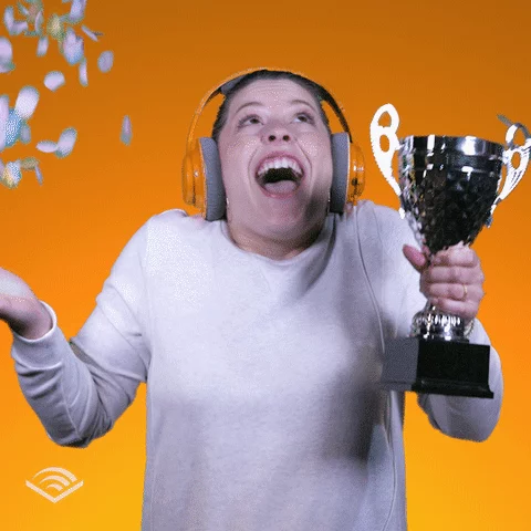 Woman wearing orange headphones looks up smiles and shakes her head while holding a trophy and confetti falls on her. 