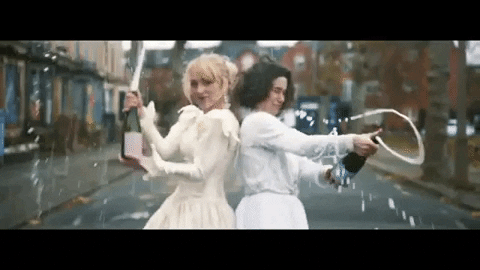 Two women in white gowns popping champagne and celebrating in the middle of the road..