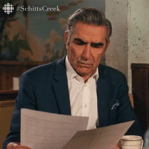 Eugene Levy wearing a white shirt and dark blazer, looking at a paper. He says, 