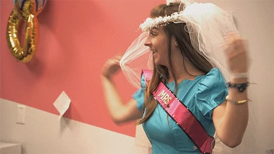 A woman dances through the office with a veil and a 'Mrs.' sash.