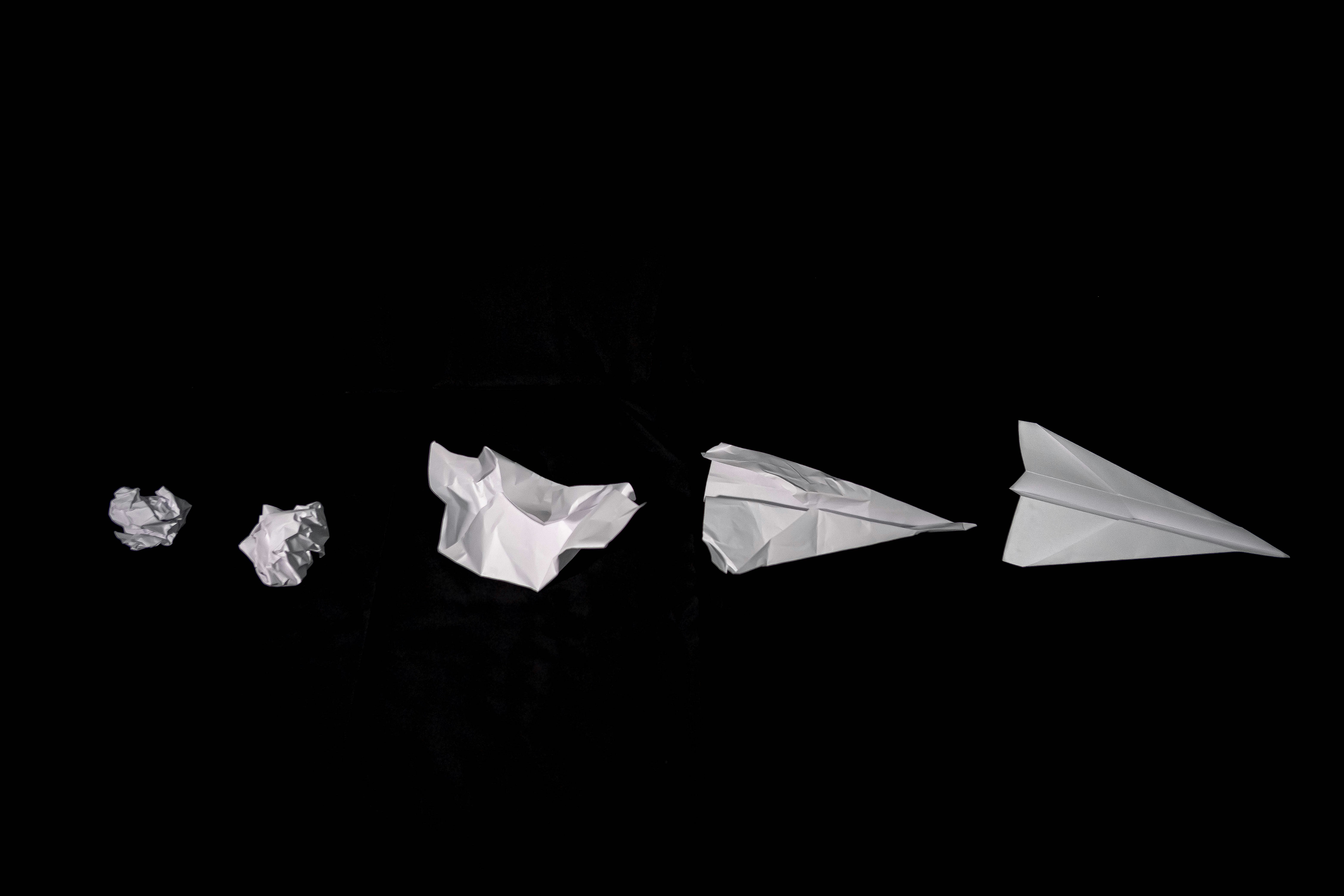 A crumpled paper gradually folding into a sleek paper airplane against a black background.