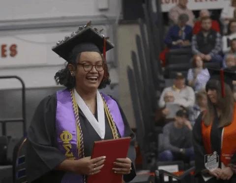 A Black graduate student receiving a diploma. She fist bumps with a staff member.