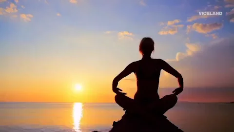 A woman doing yoga. In front of her is a view of the ocean and sunset.