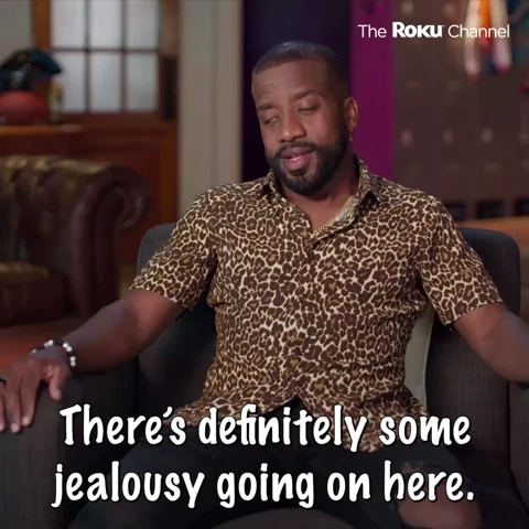 A man wearing a cheetah print shirt, saying 'There's definitely some jealousy going on here' while sitting on a brown couch