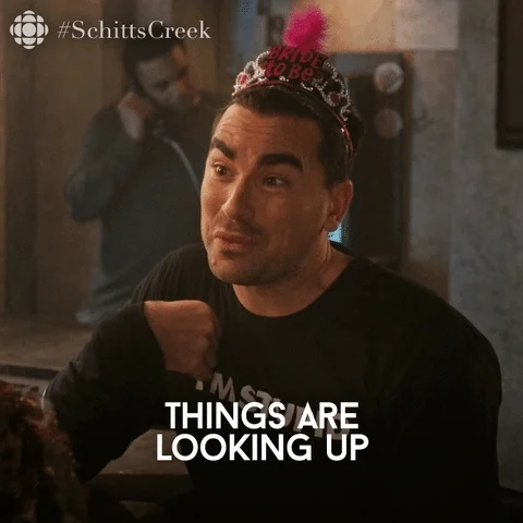 Dan Levy wearing a black long-sleeve shirt and happy new year crown with a pink feather saying, 